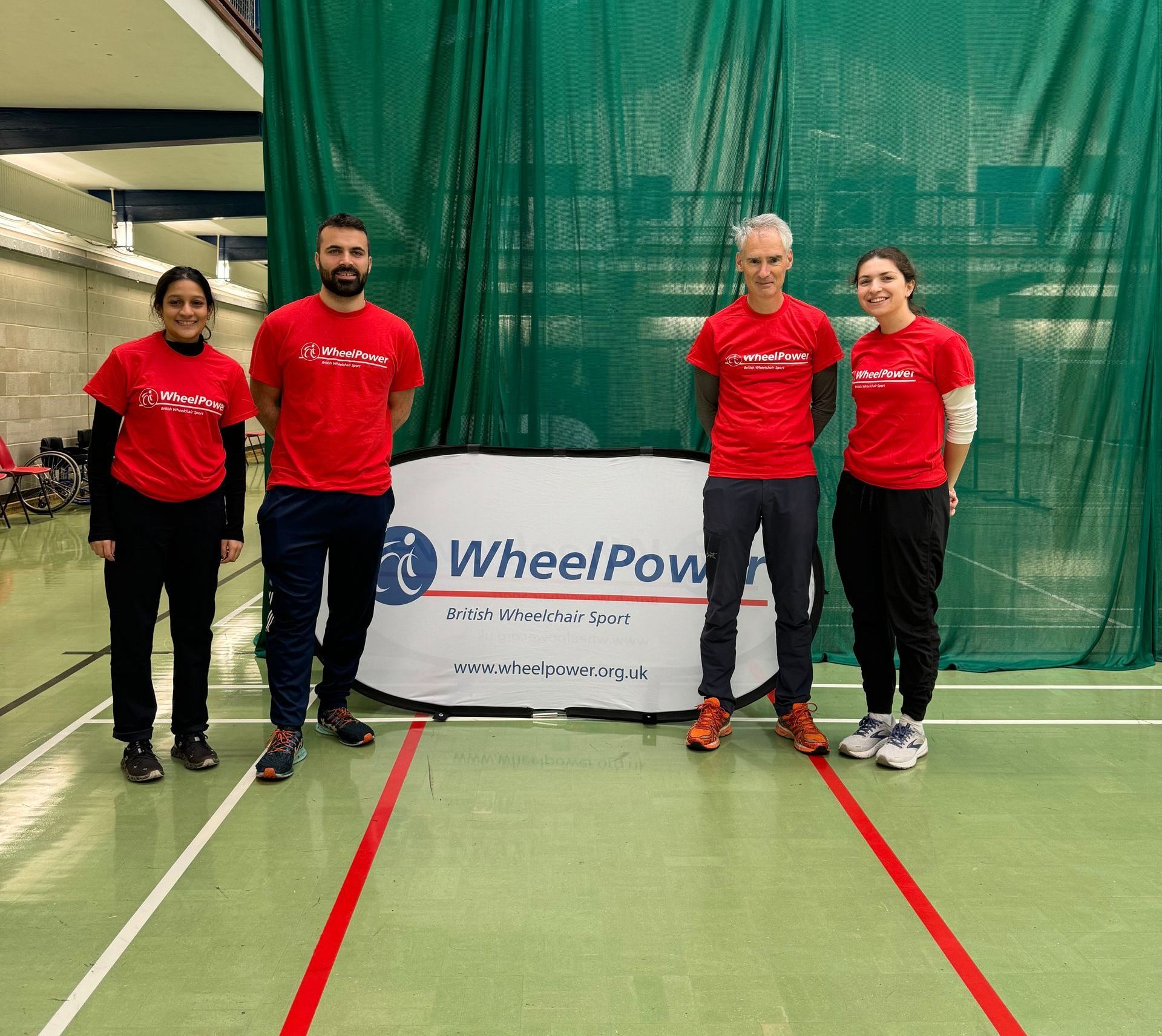 From left to right, Malavi, Charlie, Charles, and Christina stand in red Wheelpower t-shirts, with a green sports net behind and the WheelPower pop-up sign. 