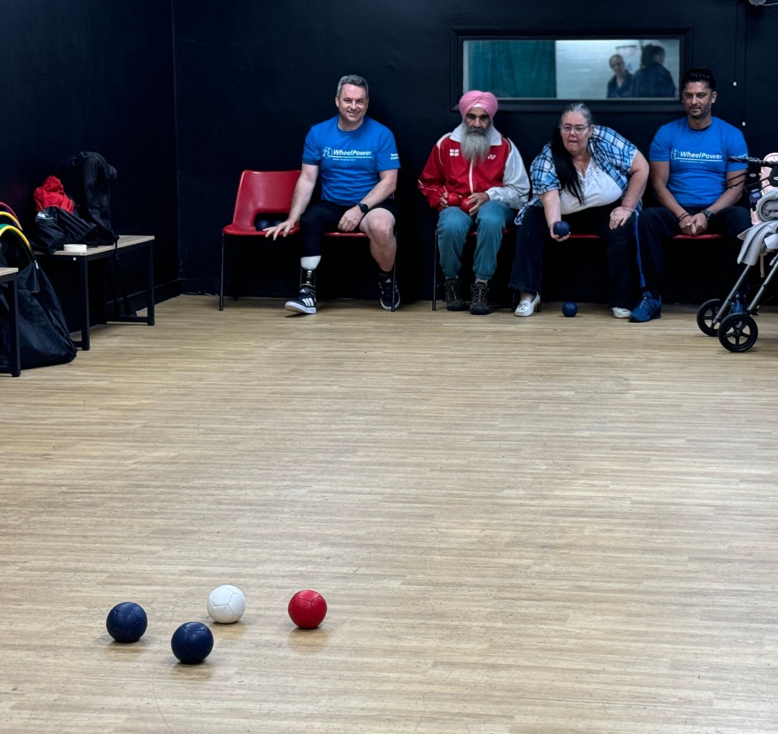 Four participants sit on a row of chairs, with boccia balls on their laps. On the wooden floor in front of them a white ball, two blue balls and a red ball sit for the game. 