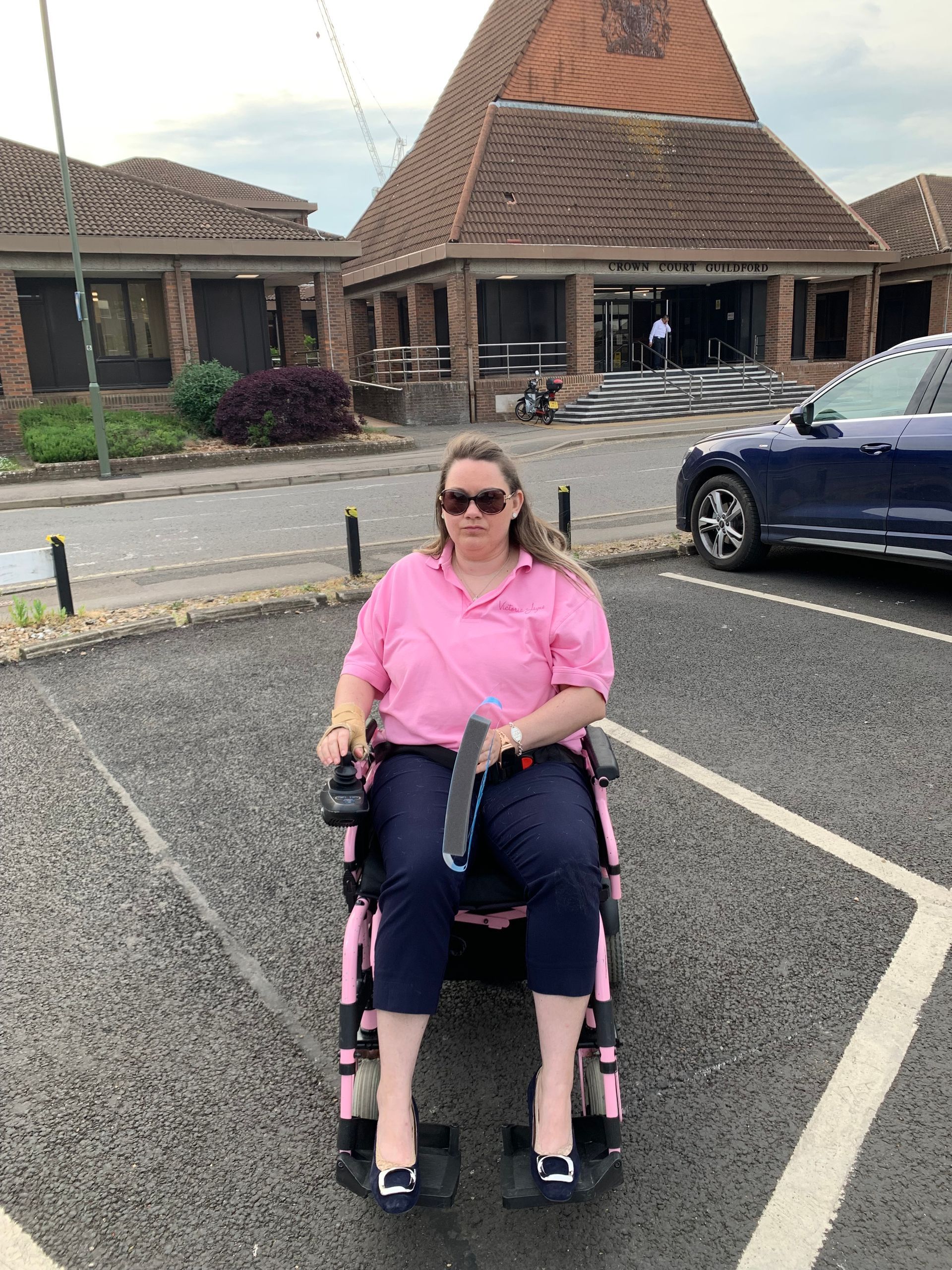 Victoria-Jayne is sat in her power chair , wearing a bright pink-shire and navy trousers. She is wearing sunglasses and has her brunette hair worn down straight. She is sat in a car park next to a car in an empty bay, with a courthouse in the background. 