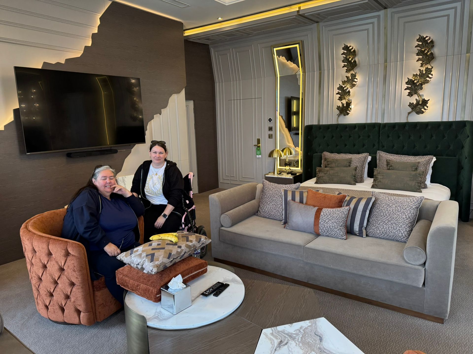 Hotel room with a grey sofa. Victoria sits in her pink wheelchair next to her mum in an armchair