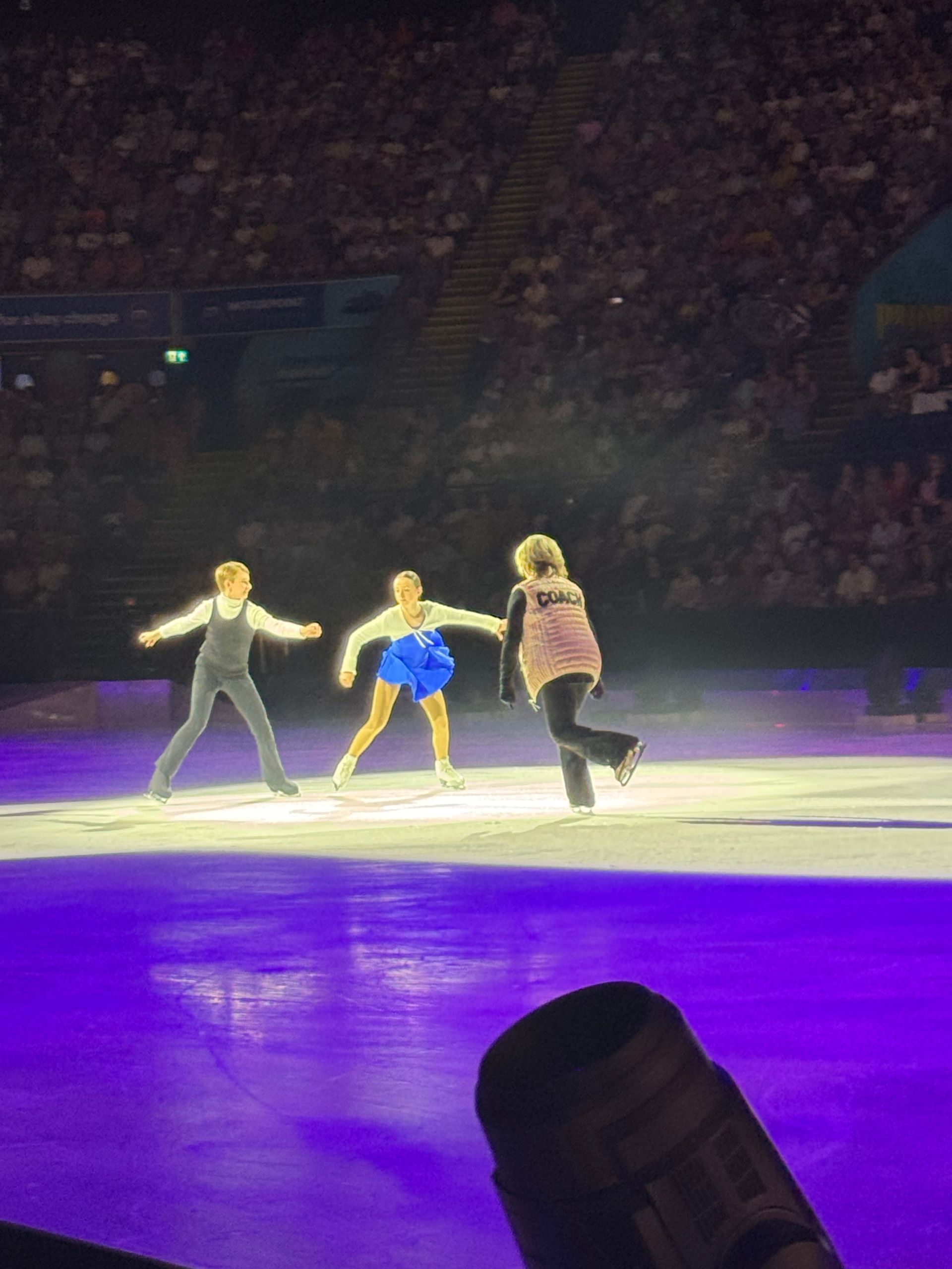 Karen Barber on the stage wearing a pink Gillet and black trousers and skates in front of a young girl wearing a blue ice skating dress and ice skates and a young lad wearing a black all in one. A spotlight in on them with the rest of the ice rink illuminated in purple. 