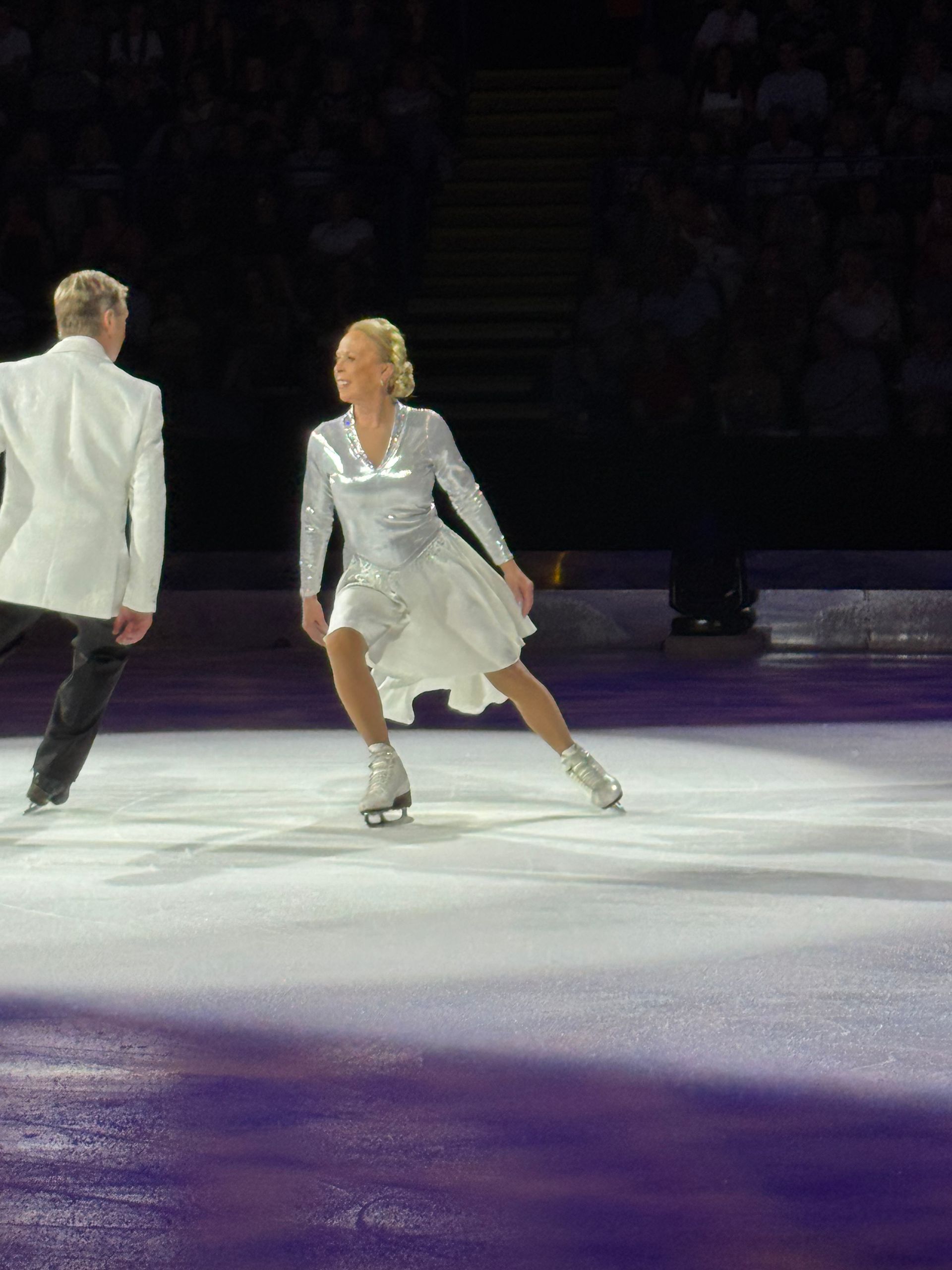 Jayne Torvill stands on the ice in a long sleeved silver top and white skirt ice skating dress, looking at Christopher Dean who is wearing black trousers and a white jacket who is looking back at Jayne as they prepare to partner to dance to Let's Face the Music. The background is in a blackout with only the pair in focus. 