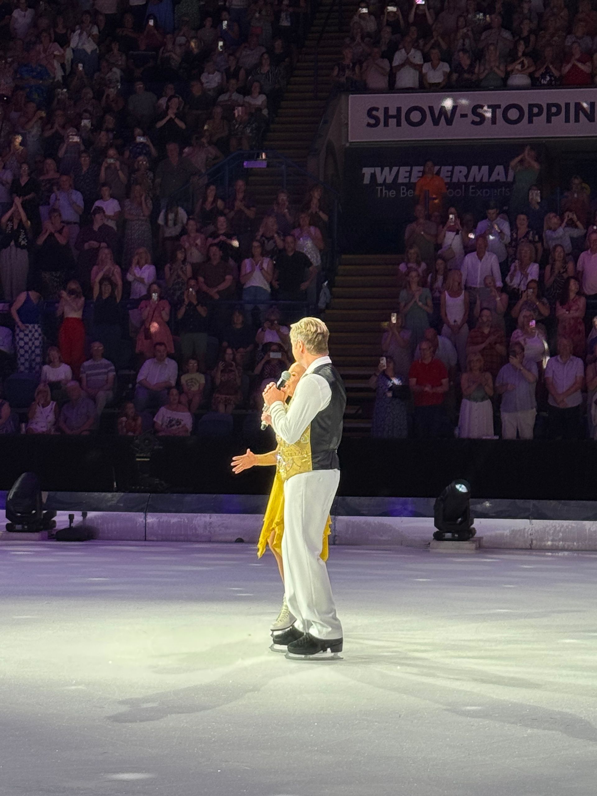 Jayne stands on the stage with a microphone in one hand and the other outstretched in gesture wearing a gold iceskating dress. 
Next to her stand Chris in white trousers white shirt and a waist coat with a black back and gold sequinned front.
The audience sit in tiered rows behind them. 