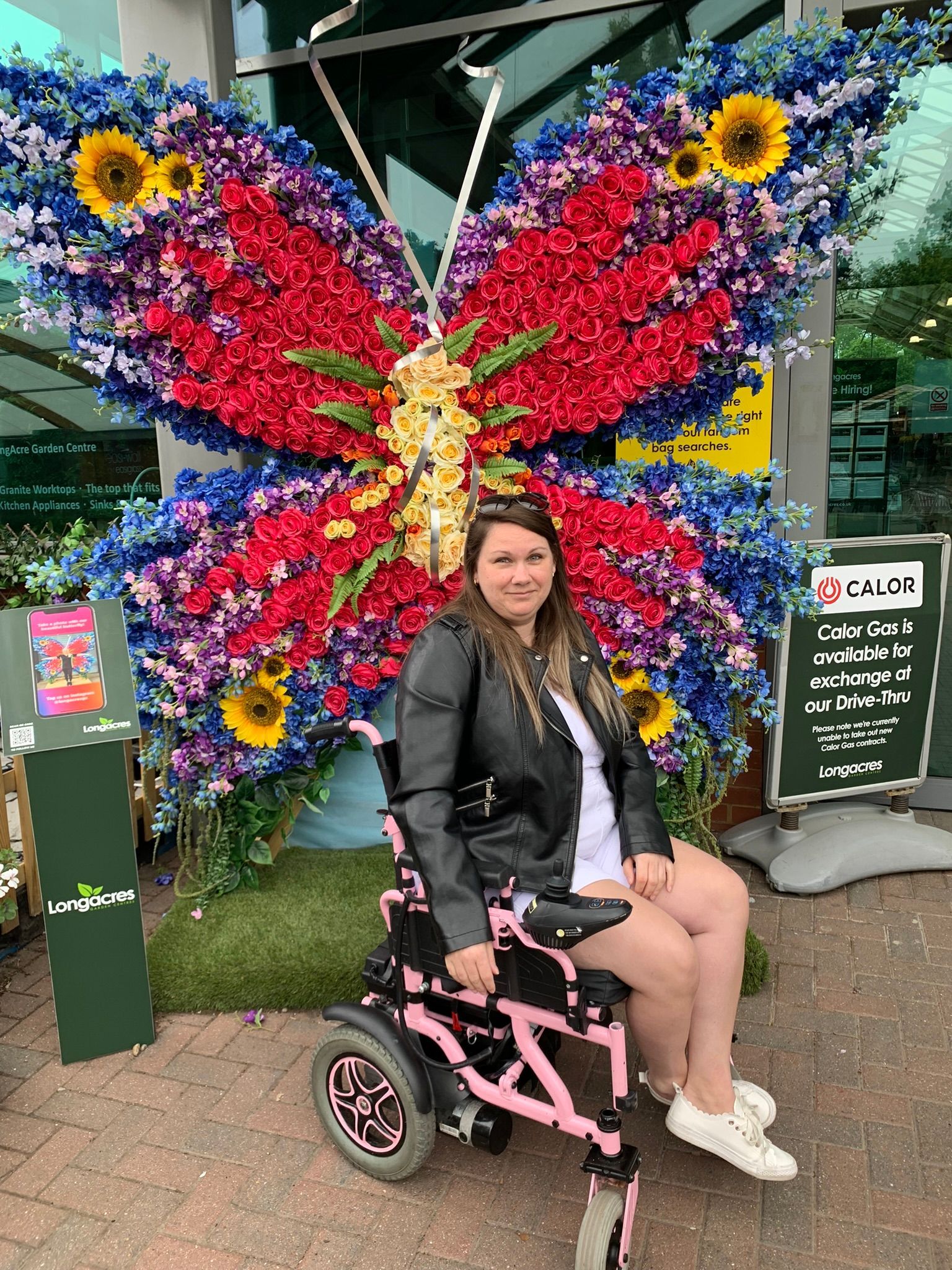 White Female with brown hair - wearing a black leather jacket and is a powerchair user which is a pink colour.
