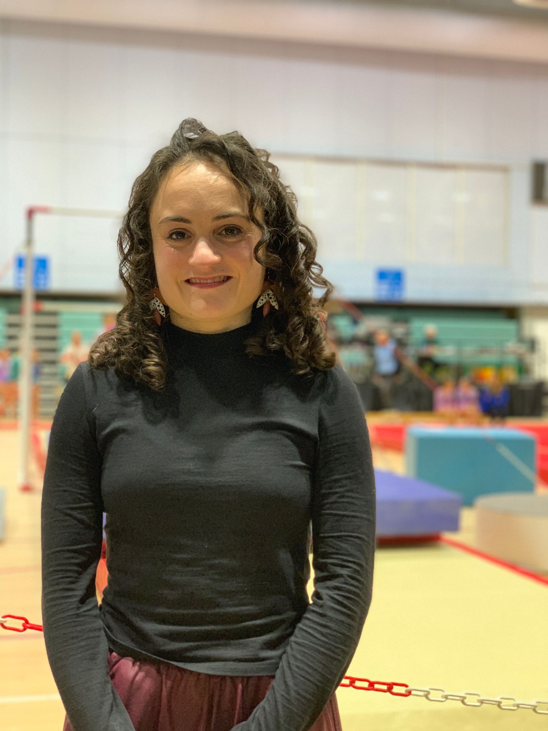 Lauren Lethbridge in a black long top with red trousers and long brown curly hair with gymnastic equipment in the background.