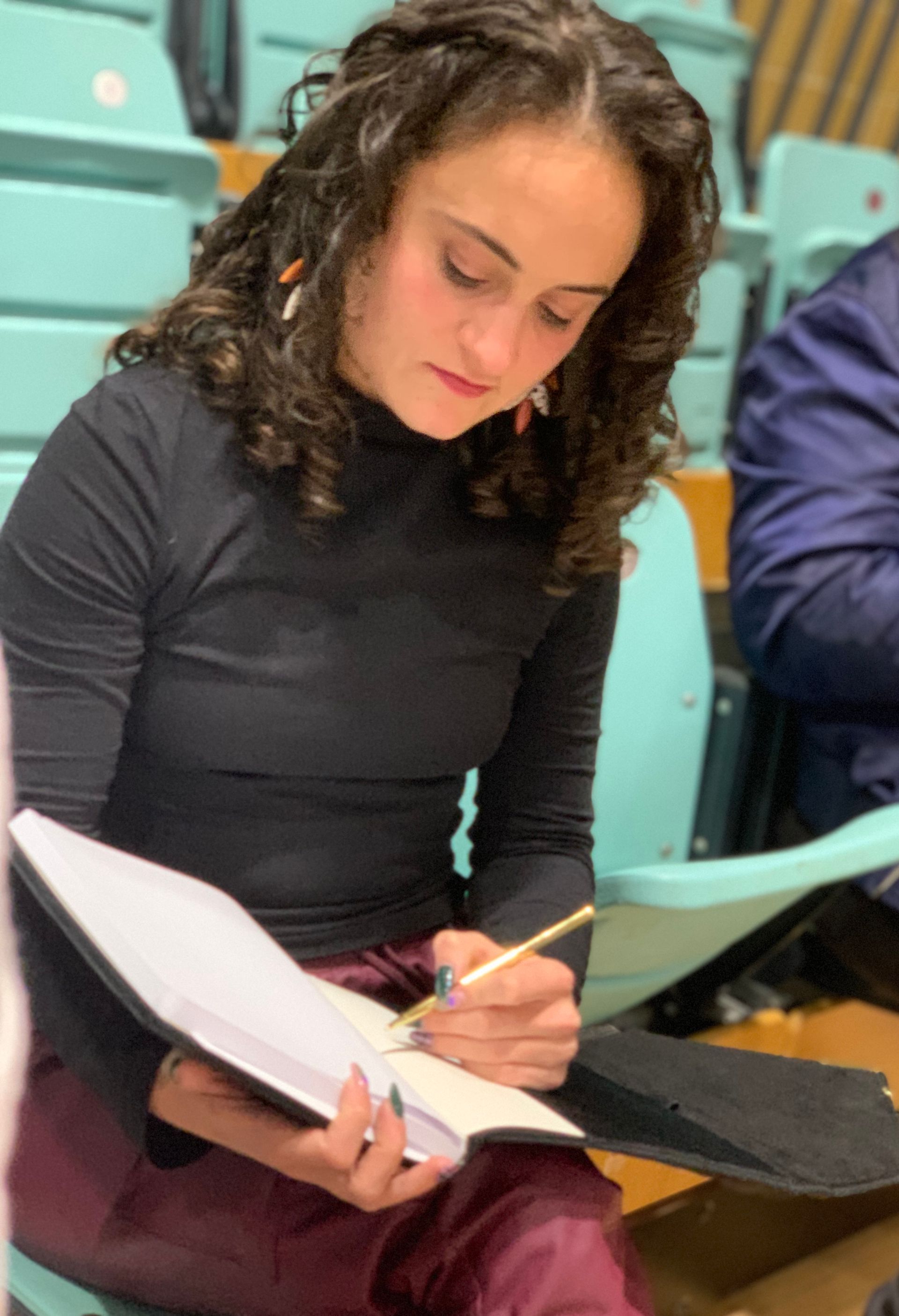 Lauren Lethbridge with long brown curly hair, in a black top with a note book open on her lap, holding a gold pen and writing notes. She is sitting on pastel green chair. 