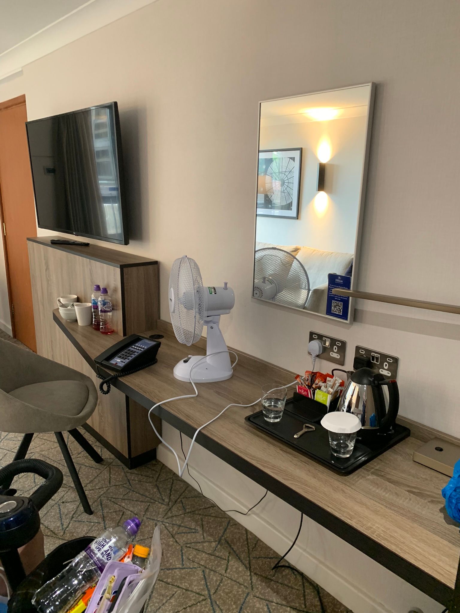 A wooden dressing table with a mirror over the top. A tray which holds a kettle and cups alongside tea and coffee sit atop the table. A TV is on the wall. 