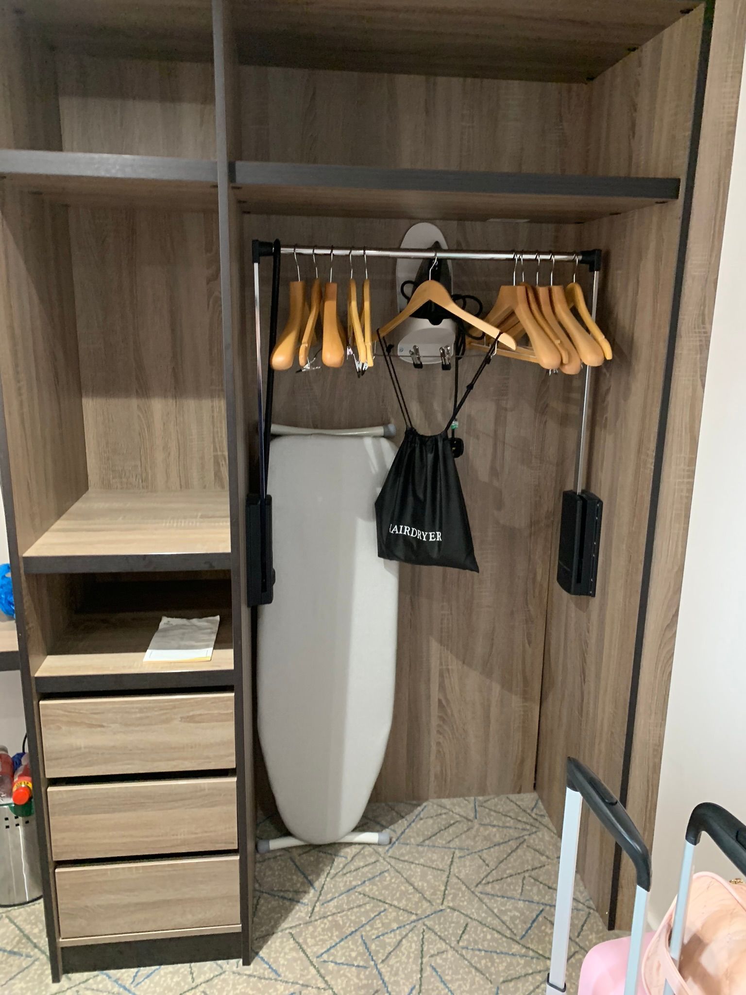 A wardrobe with a rail on the right-hand side which can be pulled down for wheelchair users. Eight wooden coat hangers sit on the rail. A white ironing board is lent up against the back of the wardrobe. To the left and side are four drawers and two shelves. 