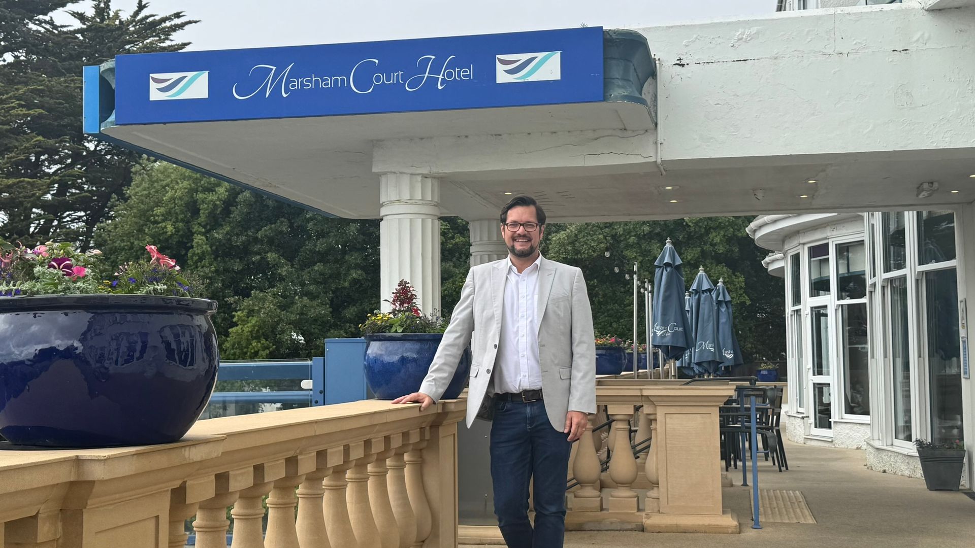 James stands with his hand on the beige wall of the terrace of the hotel. Blue flower pots with pink and green flowers line the wall. James is wearing blue jeans, a white shirt and a beige jacket. 