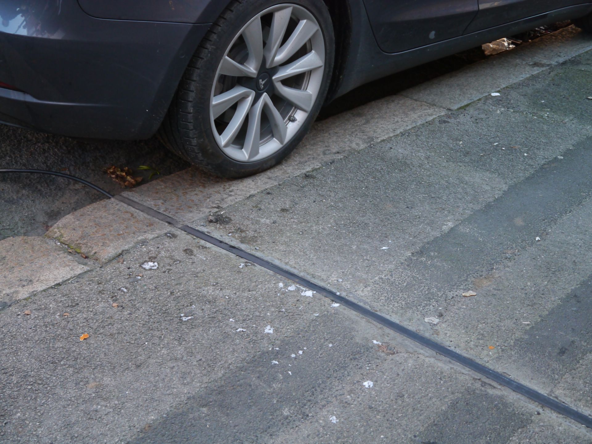 grey pavement with an electric vehicle gully cable in the middle and a blue car to the back of the picture with a wheel slightly on the pavement.  