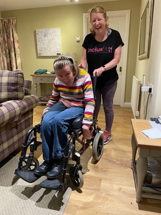 Vicki is sitting in her wheelchair, in a red and white stripped top and jeans. Sam James is stood holding onto the wheelchair wearing a black t-shirt and black leggings. 