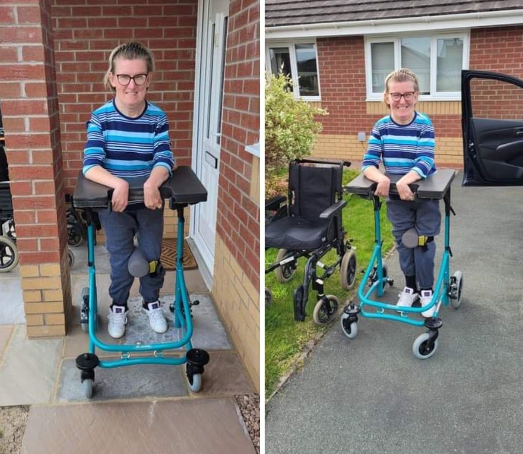 The picture is split in two. On the right, Vicki Wilson wearing a blue and black striped T-shirt is leaning on her standing walker which has black arm rests and an electric blue frame. Vicki is beaming at the camera.  On the left, Vicki is wearing the same and is leaning on her frame but is standing next to the car and has her wheelchair next to her to show she has walked from the house to car. 