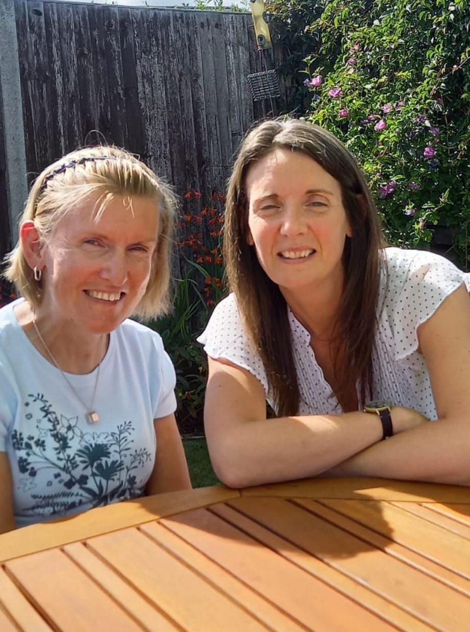 Vicki wearing a white t-shirt and squinting at the Veronica has long brown her and has a white t-shirt on. Both are sat very close to each other and leaning on a beech picnic table. 