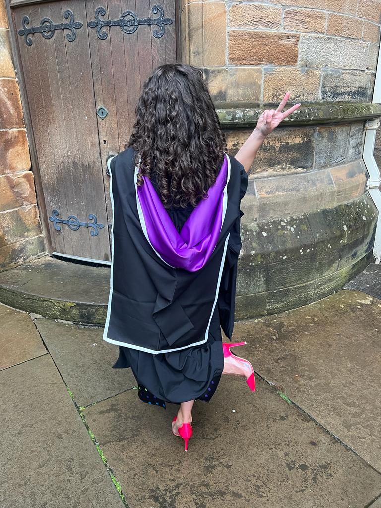 Lauren in her black graduation gown with a black and purple hood with white edgin and pink shoes. 