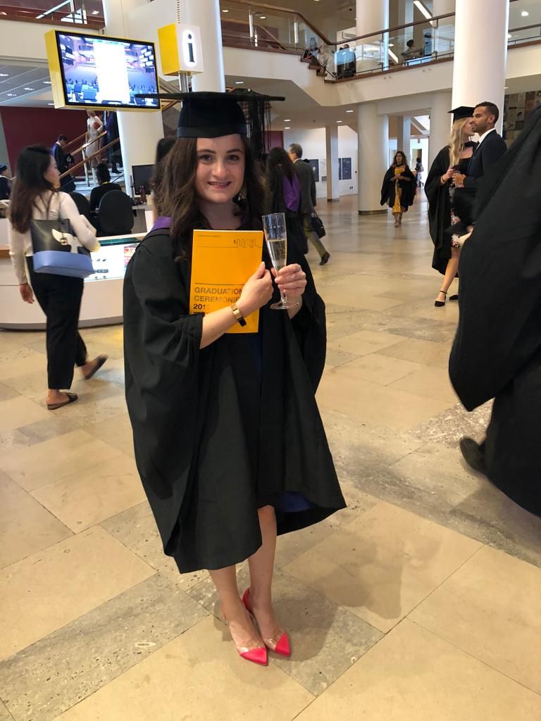 Lauren Lethbridge in a black mortarboard hat and gown, sporting a glass and champage and holding her graduation programme which is yellow.  