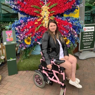Victoria sits in her pink wheelchair, wearing a leather jacket and a white t-shirt. She has long brown hair and has sunglass sat on top of her head. Behind her is a giant butterfly made of artificial flowers in reds, pinks, purple and yellow colours. 