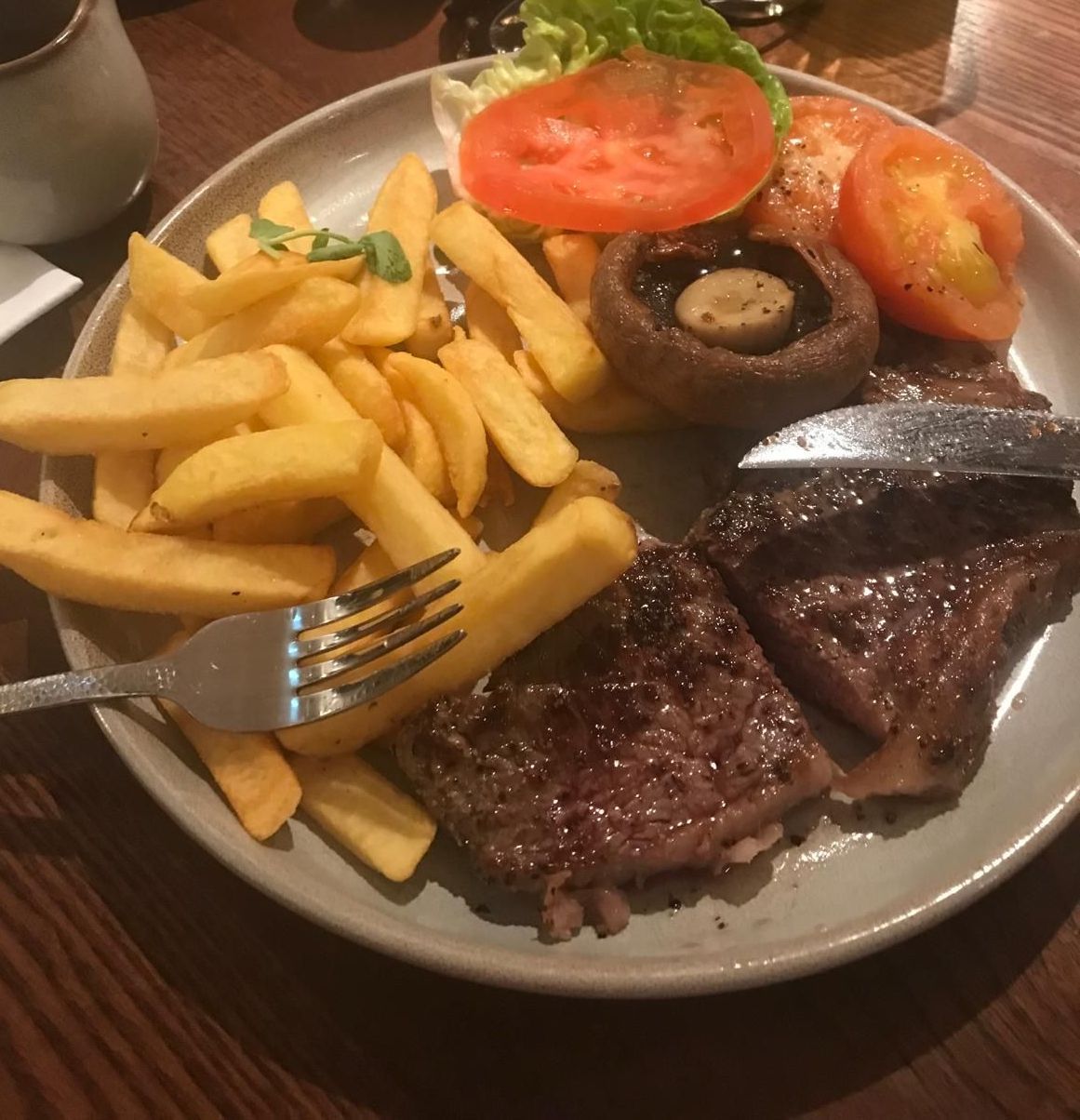 Steak, Chip, Mushroom, and tomato sit on a white plate. A brown mahogany table holds the plate. 