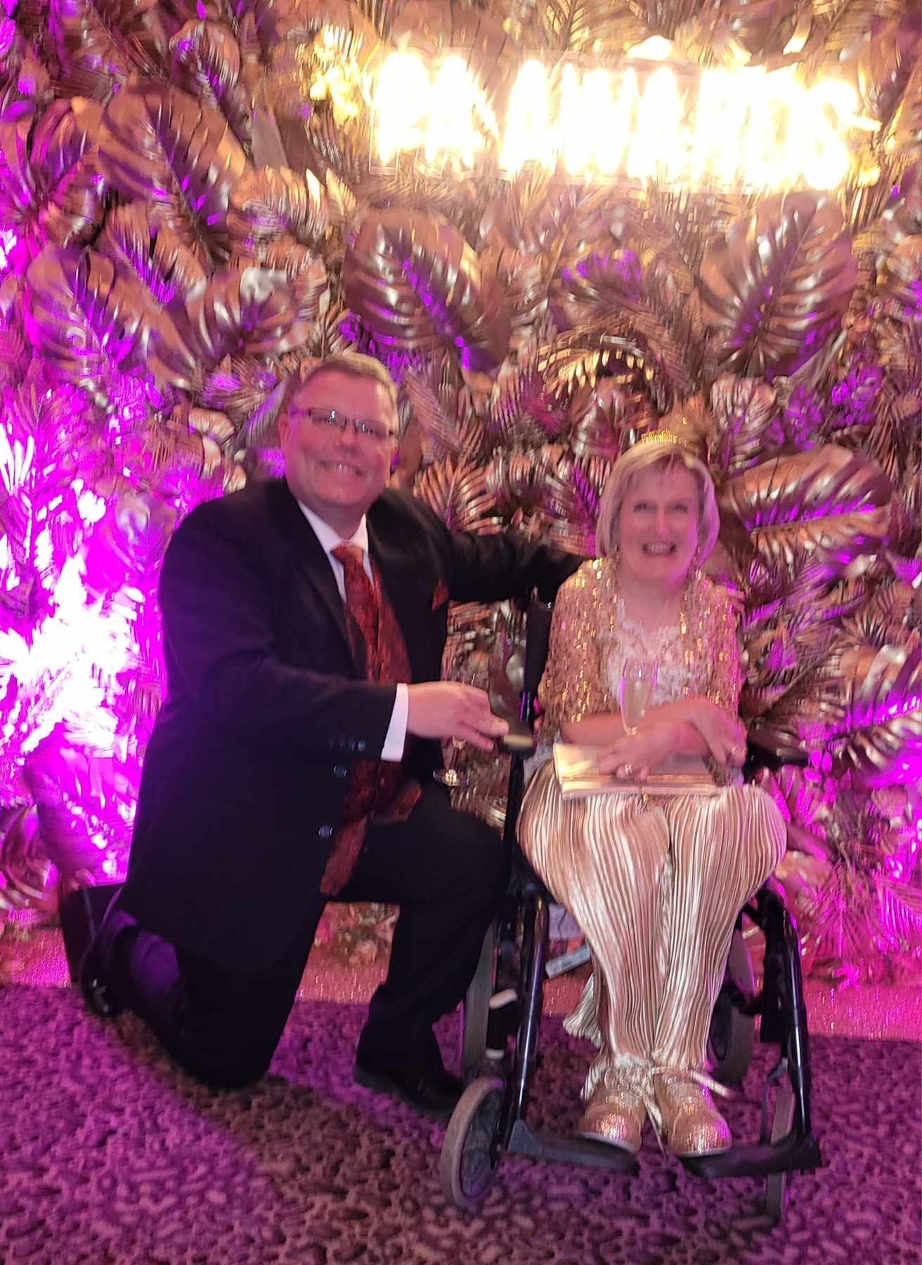 Vicki is sitting in her wheelchair wearing a glitzy gold top, gold trousers, and gold shoes. Steve is down on one knee next to her in a black suit, white shirt and red tie, on a background of gold leafs