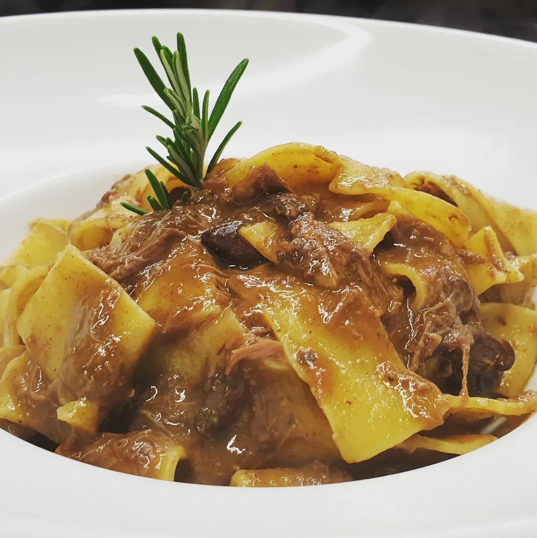 Una ciotola bianca piena di tagliatelle e carne con un rametto di rosmarino sopra