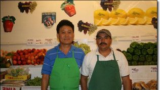 Two men are posing for a picture in front of a fruit and vegetable display.