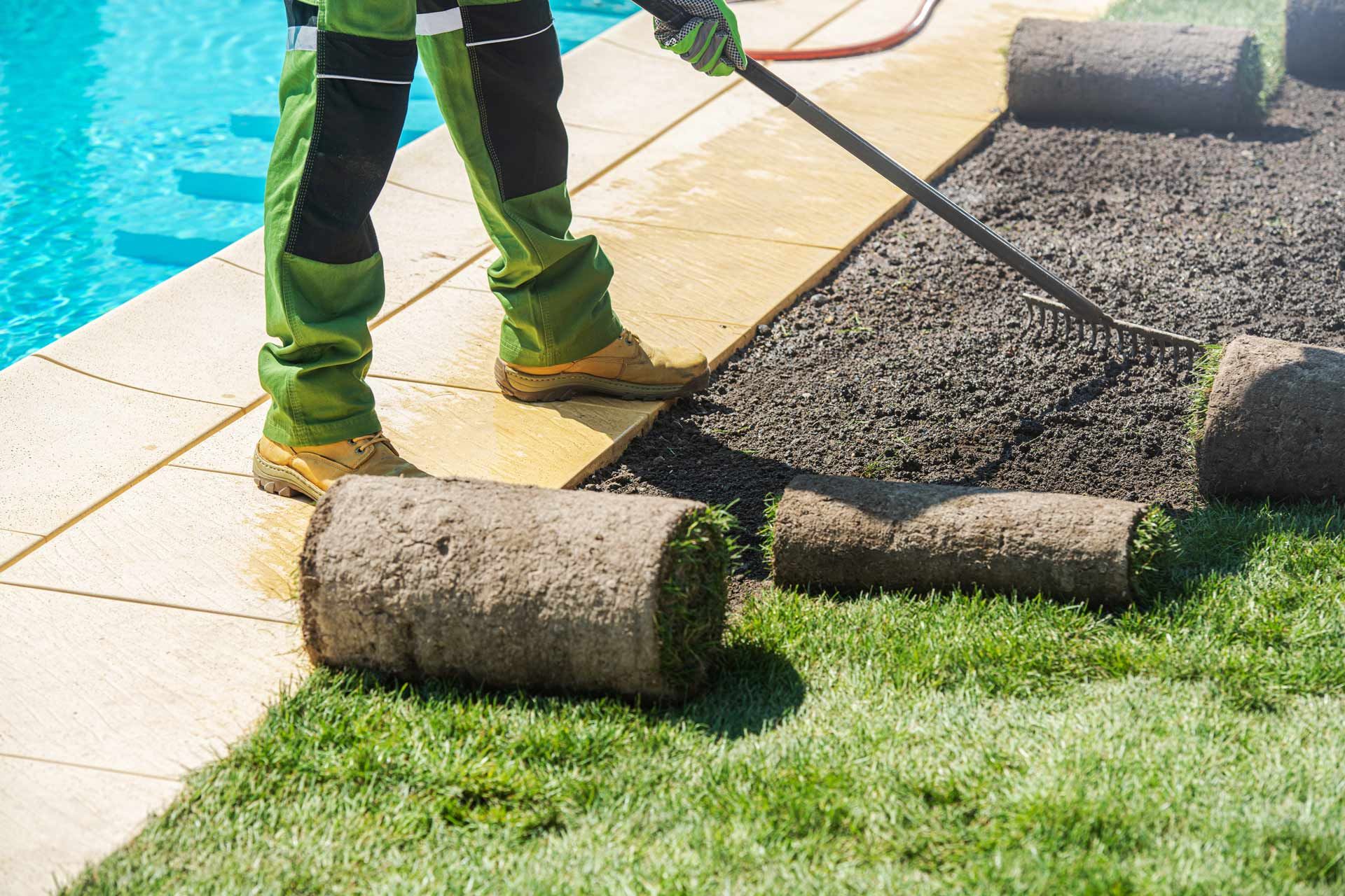Person laying sod rolls next to a swimming pool, green work clothing, gravel.