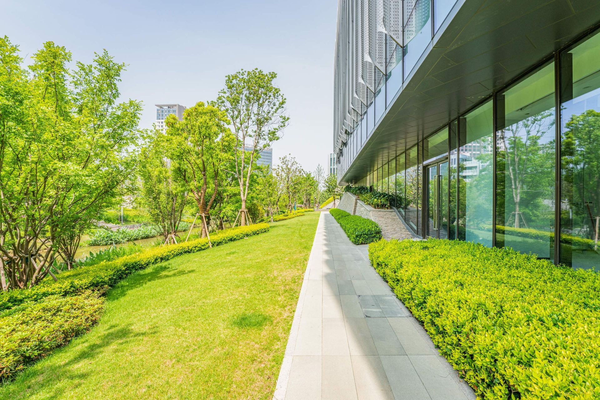 Lush green landscaping alongside a modern building with large windows. A walkway runs along the building's edge.