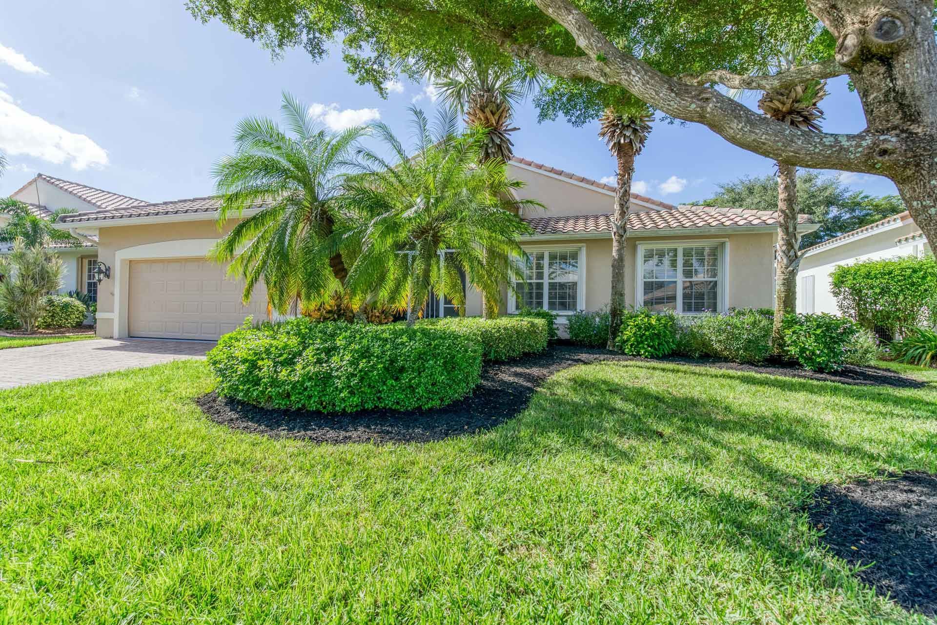 House with beige walls, tan roof, green lawn, palm trees, and bushes under a blue sky.