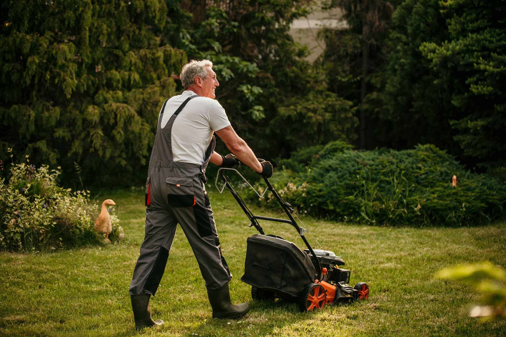 Man in overalls mowing a green lawn with a black and orange lawnmower in a yard.