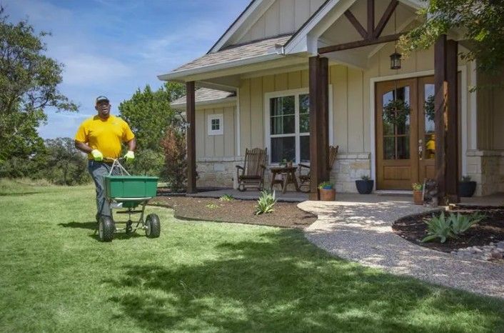 Man spreading fertilizer on lawn near a house, wearing yellow shirt and gloves.