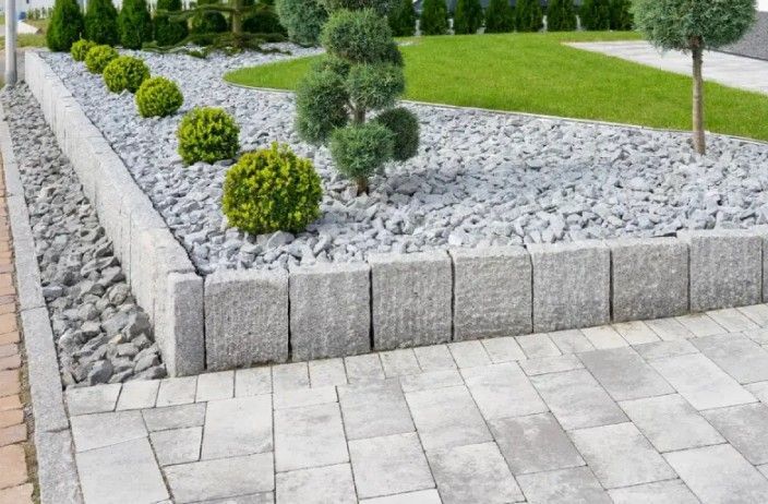 Stone-edged garden bed with gray gravel, various green shrubs, and paved pathway.