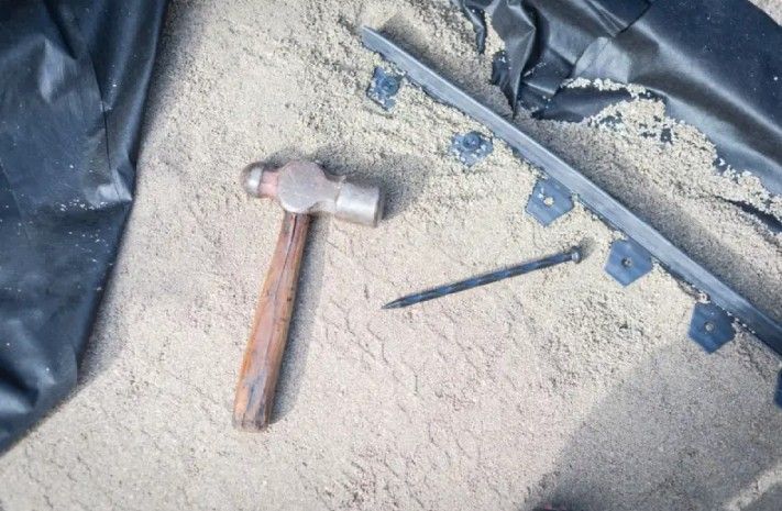 Hammer and nail on sand, next to black landscape edging and a plastic sheet.