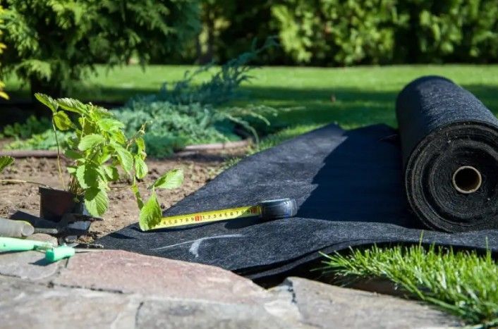 Black landscape fabric being installed in a garden bed with a measuring tape, near green plants.