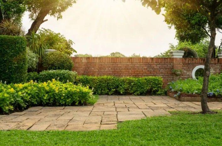 Paved patio and green lawn in a garden with brick wall, bushes, and trees in sunlight.