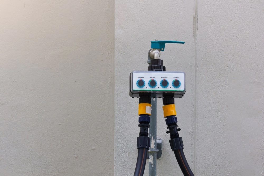 Water faucet with a 4-zone timer controller and hoses attached against a light-colored wall.