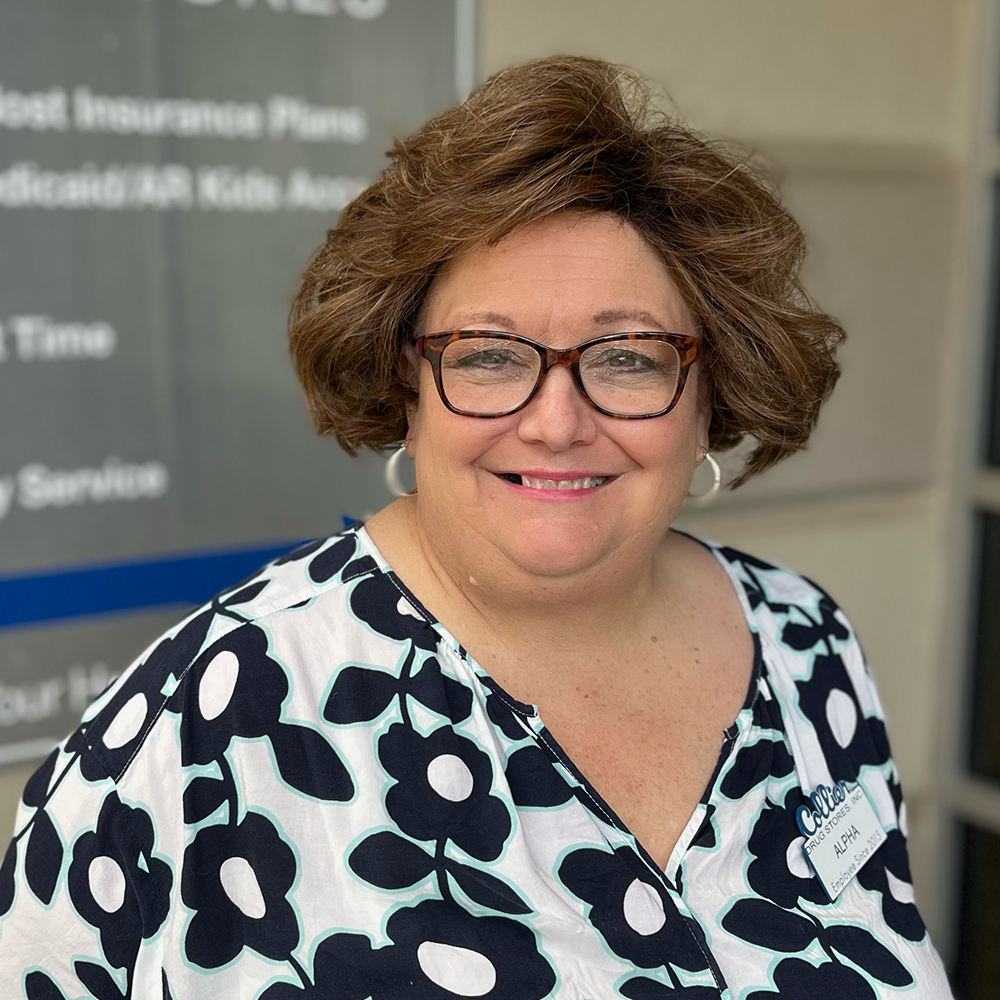 A man in a blue scrub is smiling in front of a pharmacy.
