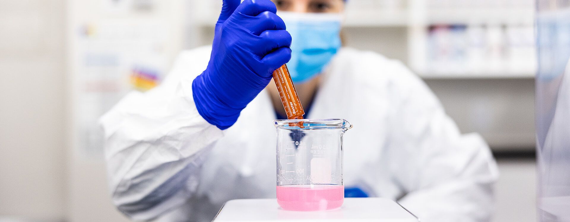 A scientist wearing a mask and blue gloves is pouring liquid into a beaker.