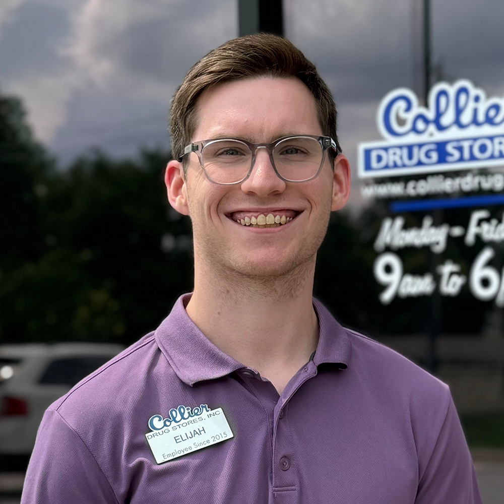 A young man wearing glasses and a blue scrub is standing in front of a pharmacy.