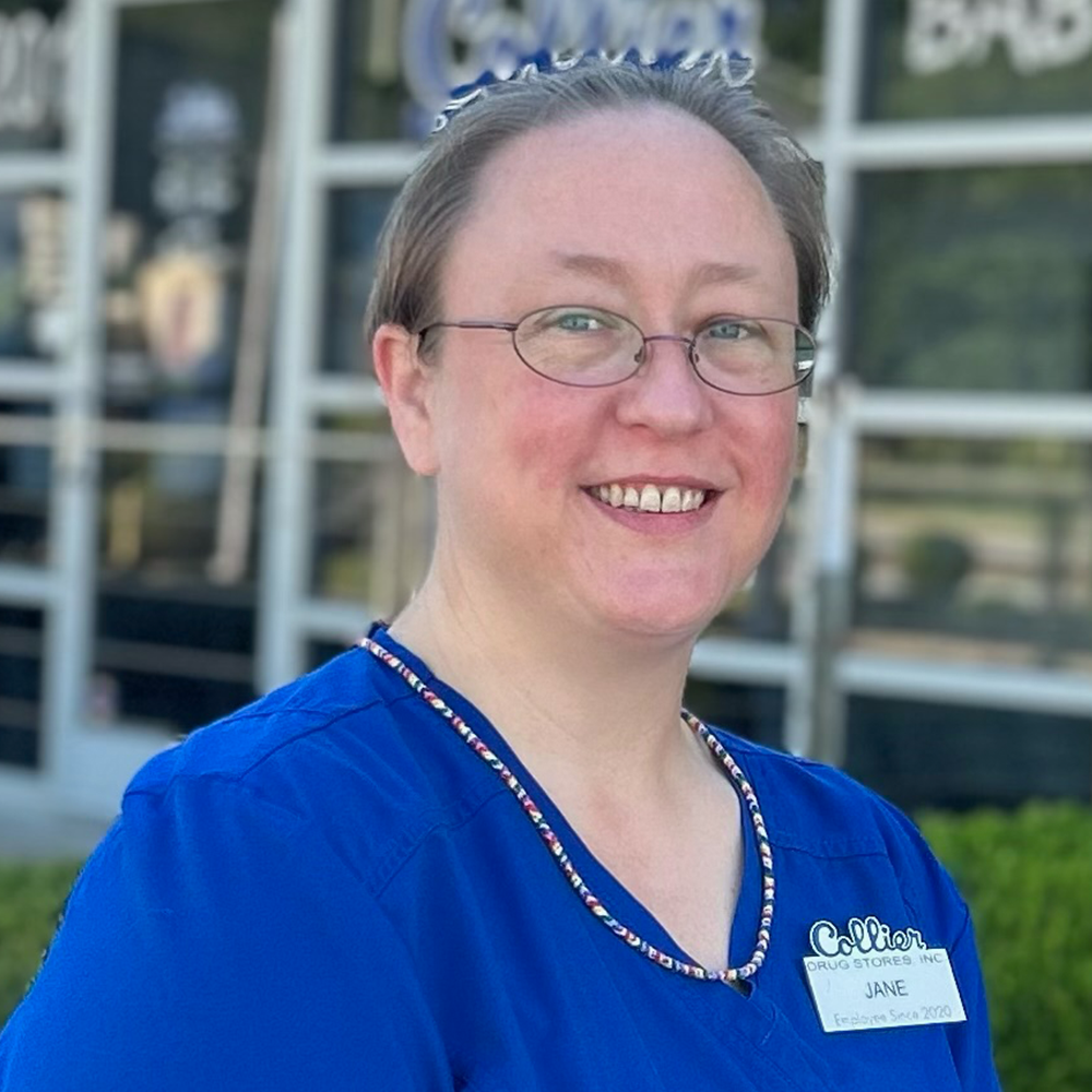 A young man wearing glasses and a blue scrub is standing in front of a pharmacy.