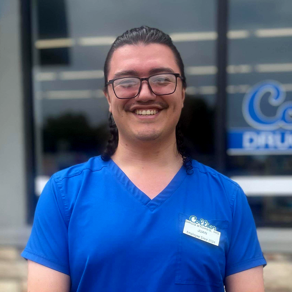 A man wearing glasses and a blue scrub top is smiling in front of a building.