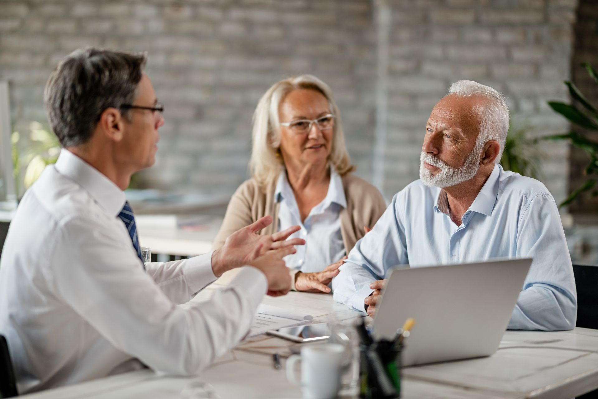 A professional meeting between an advisor and two individuals seated at a table with a laptop in a brick-walled office.
