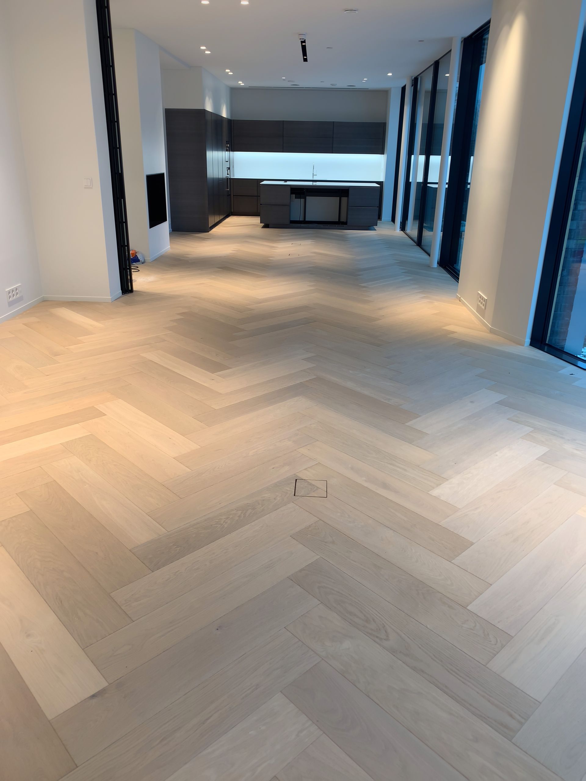 Et stort tomt rom med fiskebeinsgulv og kjøkken i bakgrunnen.