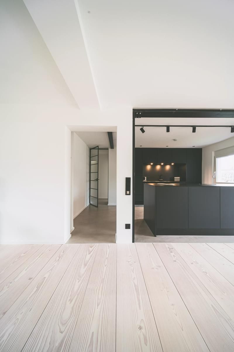 Et tomt rom med tregulv og kjøkken i bakgrunnen.