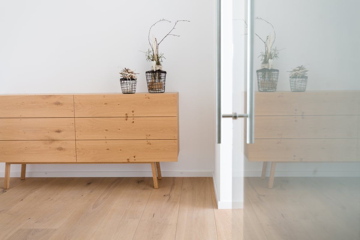 En kommode i tre med to potteplanter på står i et rom ved siden av en glassdør.