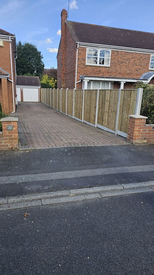 A wooden fence surrounds a driveway leading to a brick house.