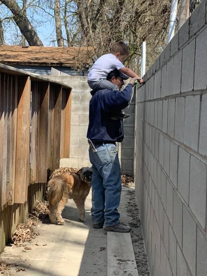 A man is carrying a child on his shoulders while a dog watches.