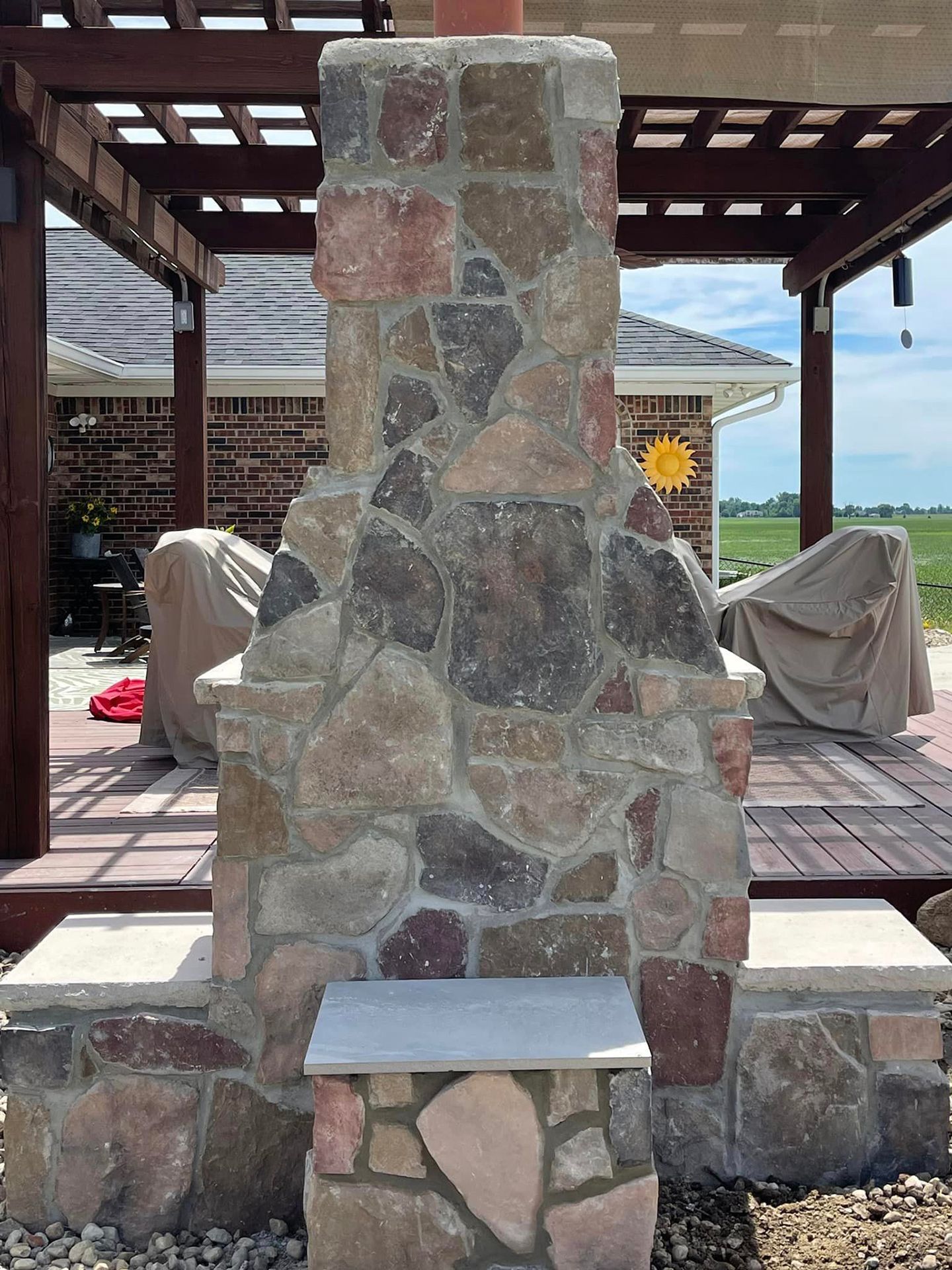 A stone fireplace is sitting under a pergola on a patio next to a house.