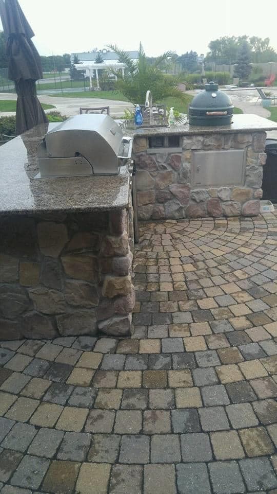 An outdoor kitchen with a grill, sink, and umbrella on a brick patio.