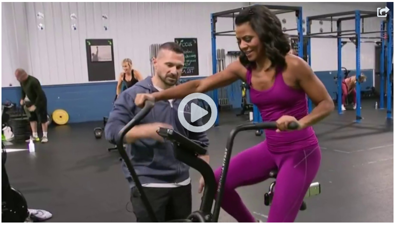 A woman is riding an exercise bike in a gym while a man watches.