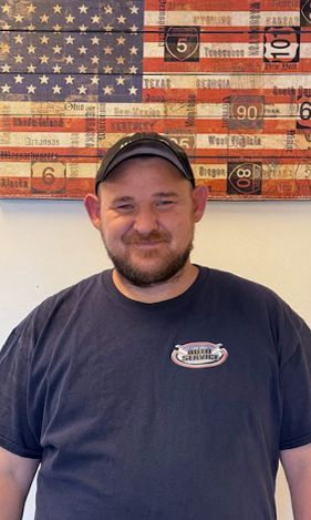 Man wearing a baseball cap and blue t-shirt with logo, smiling in front of an American flag display.