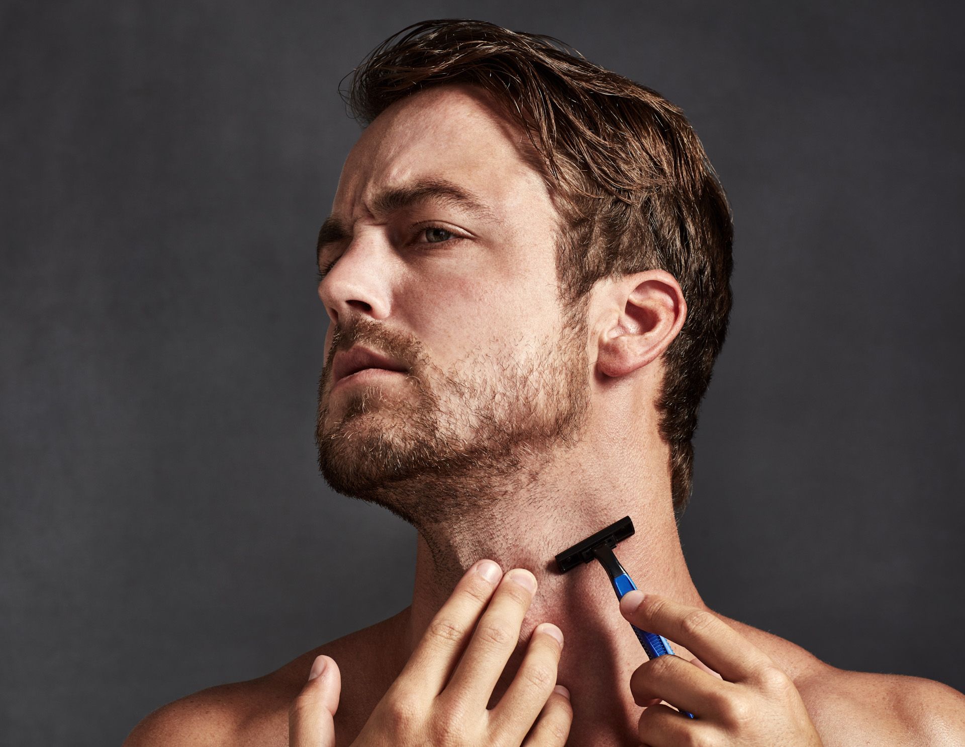 A shirtless man is shaving his neck with a razor.